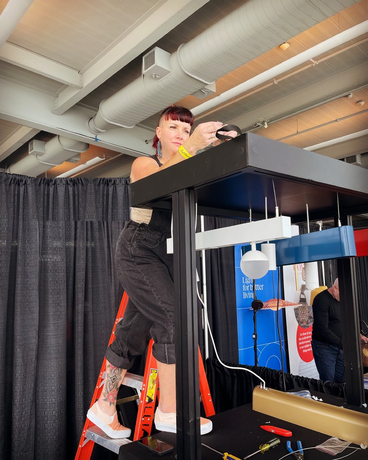 Micaila on a ladder installing a linear pendant at a lighting showcase booth