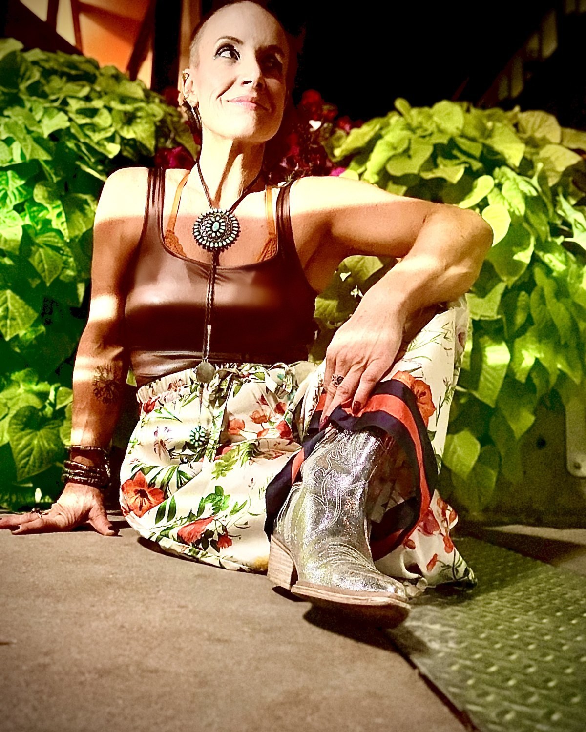 Micaila seated among lit greenery in a floral skirt, bolo tie, and silver boots